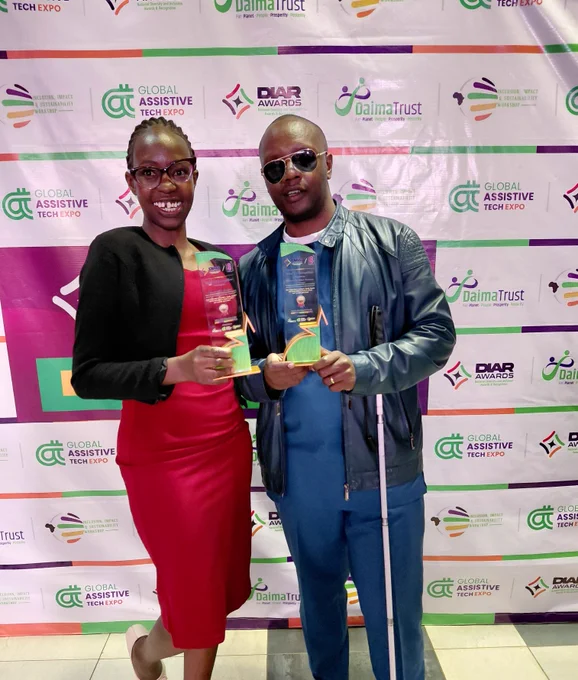 Two people stand side by side in front of a branded event backdrop featuring logos for the DIAR Awards, Daima Trust, and the Global Assistive Tech Expo. They are smiling and each holding a glass award trophy.

The person on the left is wearing a red dress with a black blazer and glasses, while the person on the right is dressed in a blue outfit with a black leather jacket, sunglasses, and holding a white cane. The setting appears to be an awards event celebrating diversity and inclusion.