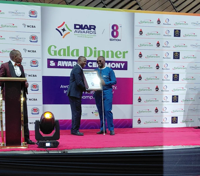 A man stands on a stage at the DIAR Awards 8th Edition Gala Dinner and Award Ceremony, receiving a framed certificate from another man in a suit. He is dressed in a blue outfit, wearing sunglasses, and holding a white cane.

They are smiling as they shake hands during the presentation. The stage has a red carpet, a podium to the side, and a branded backdrop featuring DIAR Awards signage and sponsor logos including Safaricom, KPC, and KenGen.