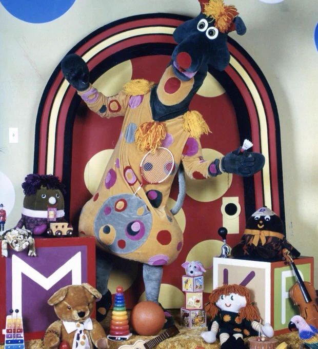 The Polkaroo standing in a colorful children’s television set from *Polka Dot Door*. The tall, kangaroo-like character has a long dark head with large round eyes, a red nose, and a tuft of yellow-orange hair. It wears a tan costume covered in large multicolored polka dots with tassels on the shoulders and holds a badminton shuttlecock in one hand. Behind it is a red arch doorway with large yellow polka dots. Surrounding the character are numerous toys and props, including alphabet blocks with the letters “M,” “K,” and “R,” stuffed animals, dolls, stacking rings, a small guitar, a toy telephone, a drum, a violin, a rubber duck, and other children’s play items arranged across the floor.
