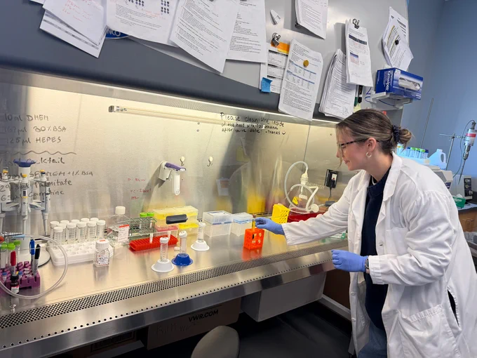 A person wearing a lab coat and gloves is working at a laboratory bench filled with various scientific equipment and labeled containers. Notes and papers are attached to the wall above the bench.