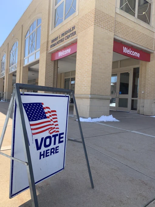 Sign with an American flag reading 'VOTE HERE' stands outside Edgewood University Predolin Hall's entrance with 'Welcome' banners.