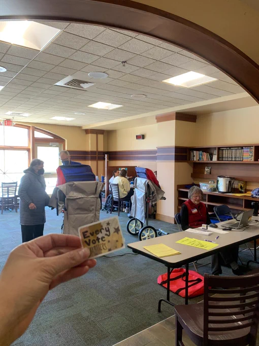 Hand holding a voting sticker reading 'Every Vote Matters' inside an in-person absentee voting site at Edgewood University Common's  with poll workers helping with voters and interacting at tables.