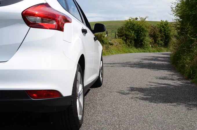 Rear side view of a white car parked on a quiet country road with greenery and hills in the background.