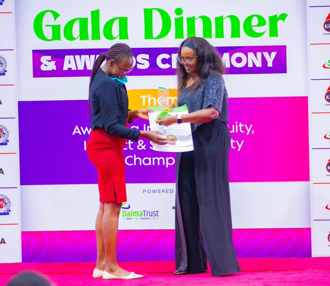 A photograph taken on stage during the DIAR Awards Gala Dinner & Awards Ceremony.

Two women are standing on a red carpet in front of a colorful purple, white, and pink backdrop. The woman on the left, wearing a black blazer and a bright red skirt, is officially presenting the EnableMe Kenya certificate and glass trophy to the woman on the right. The recipient is wearing a shimmering dark grey top and black wide-leg trousers, smiling as she accepts the award.

The backdrop prominently features the text "Gala Dinner & Awards Ceremony" and the DaimaTrust logo at the bottom. The atmosphere is celebratory and professional, capturing the moment of recognition for the organization's work in diversity and inclusion.