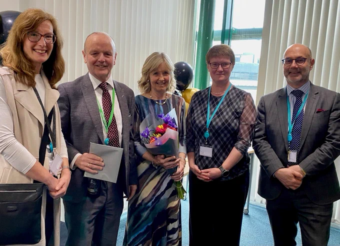 Five people standing together. One is holding a document. One is holding a bunch of flowers.
