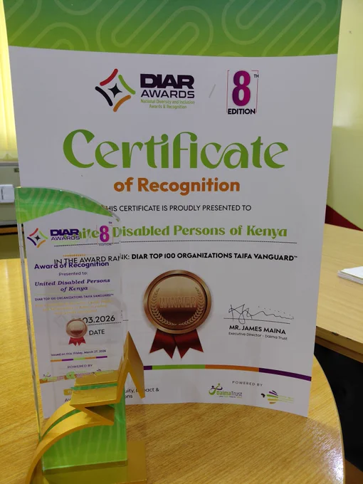 A Certificate of Recognition and a glass trophy awarded to the United Disabled Persons of Kenya (UDPK) at the 8th Edition of the DIAR Awards (National Diversity and Inclusion Awards & Recognition).

The certificate features a vibrant green and white wavy pattern at the top. It identifies the organization's rank as "DIAR TOP 100 ORGANIZATIONS TAIFA VANGUARD" and includes a gold "Winner" seal with a red ribbon. The document is signed by Mr. James Maina, Executive Director of Daima Trust, and is dated March 2026.

In the foreground, a matching tall acrylic trophy with green and gold accents stands on a light wooden table. The trophy's base is shaped like a stylized star or "M," reflecting the award's branding.