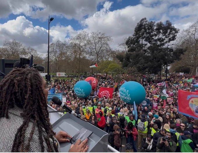 Bell Ribeiro-Addy, MP for Clapham & Brixton Hill, speaks at a podium in a park, addressing a large rally. A dense crowd stretches far into the distance, holding colourful union banners, flags, and balloons, including Unite and National Education Union branding. Several stewards in high‑visibility vests stand near the front, with leafless trees and a partly cloudy sky in the background.