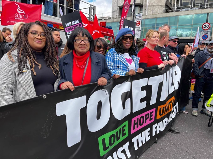 A group of demonstrators, including Bell Ribeiro-Addy, Diane Abbott, Dawn Butler and Sharon Graham, stand together on a city street holding a large black banner that reads “TOGETHER LOVE HOPE UNITY AGAINST THE FAR RIGHT.” Behind them are red Unite the Union and TUC flags. Buildings, a bus, and a 20 mph “Controlled Zone” sign are visible in the background.