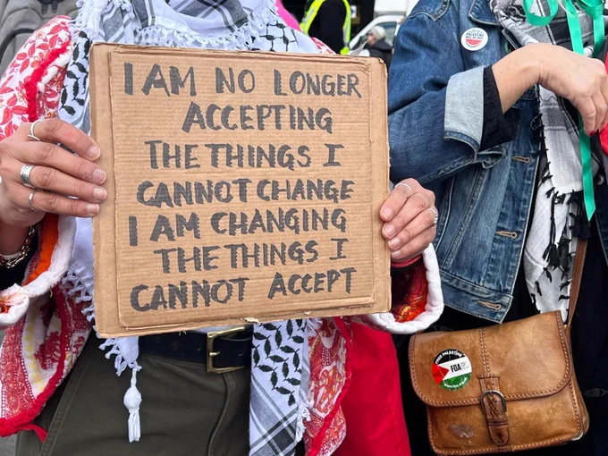 Two people stand close together at a public demonstration. One holds a cardboard sign reading “I am no longer accepting the things I cannot change. I am changing the things I cannot accept.” They wear a patterned scarf and rings. The person beside them wears a denim jacket and carries a brown leather bag with a circular badge featuring the Palestinian flag colours. Other attendees and high‑visibility vests are visible in the background.