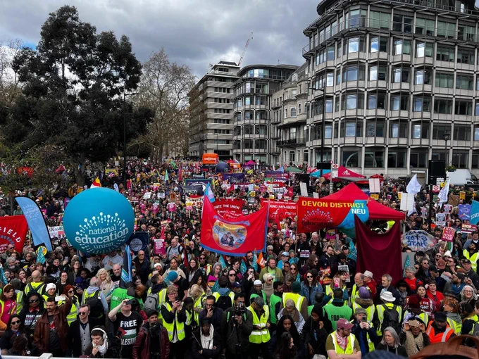 A large crowd fills an urban street during a public rally. People hold colourful union banners and flags. High‑visibility vests are scattered throughout the crowd, with modern buildings and trees surrounding the scene.