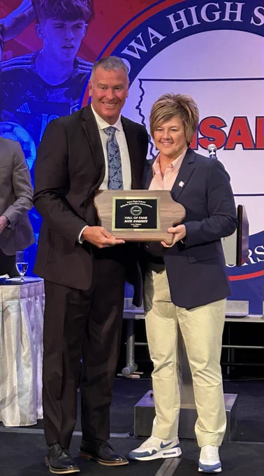 Two individuals stand on stage holding a Hall of Fame award plaque at a formal event with a school emblem backdrop.
