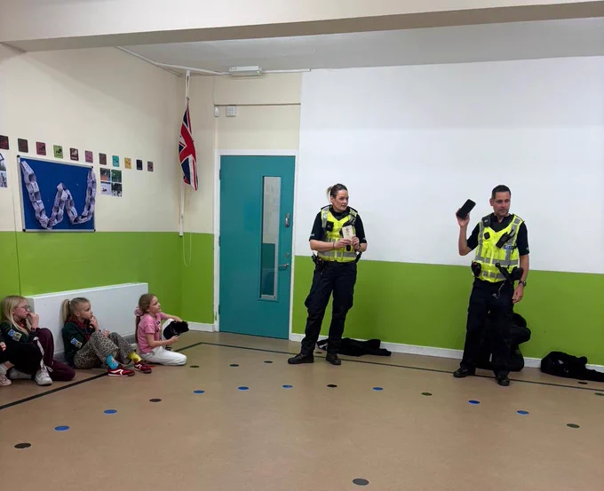 Two uniformed police officers standing in a hall giving a talk to a group of children sitting down