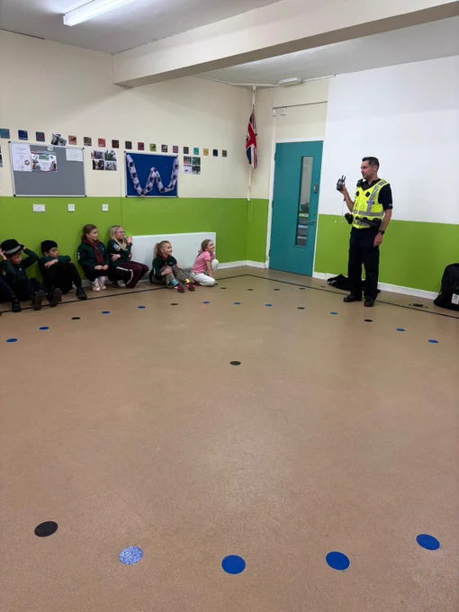 Two uniformed police officers standing in a hall giving a talk to a group of children sitting down
