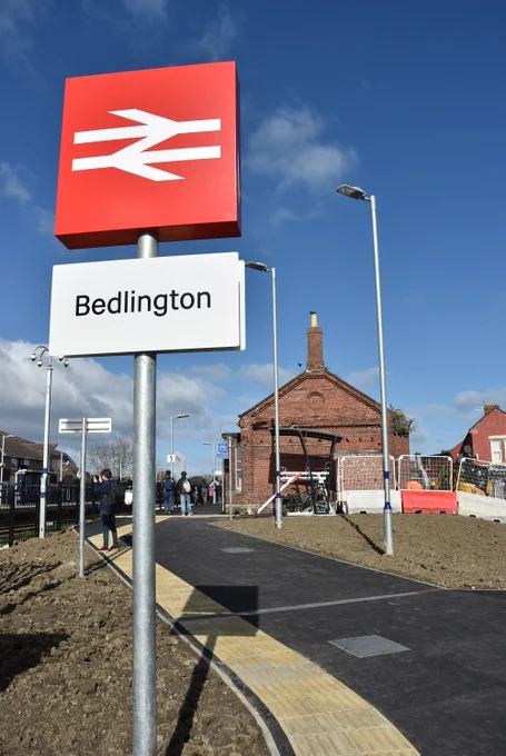 Bedlington station double arrow sign at corner of Ravensworth St and Clayton Road with blue sky behind it on a sunny day. Old building in middle distance from previous station is to be repurposed into a restaurant and tarmac path leading up to Newcastle bound platform.