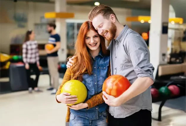 A young woman with red hair and a man with a beard smile as they stand together in a bowling alley, each holding a bowling ball; the woman holds a yellow ball and wears a denim shirt with an orange cardigan, while the man holds an orange ball and wears a grey button-down; in the blurred background, other people are visible near the bowling lanes.