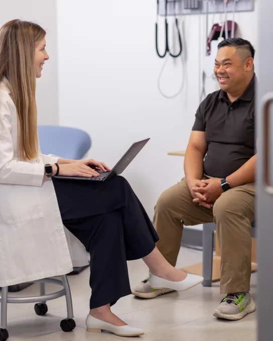 A person in a white coat with a laptop is sitting on a stool, smiling at another person seated across from them. They are in a medical office setting.