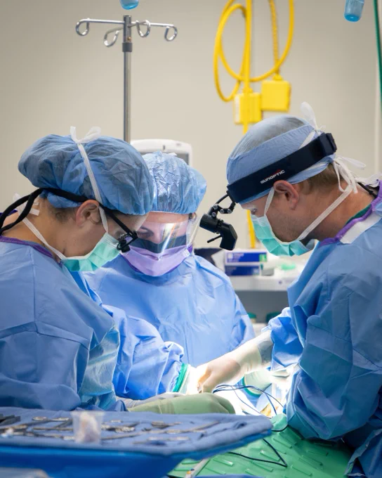 Three medical professionals dressed in blue surgical attire are performing surgery in a well-equipped operating room. They are focused on their task, with various medical instruments and equipment visible in the foreground and background.