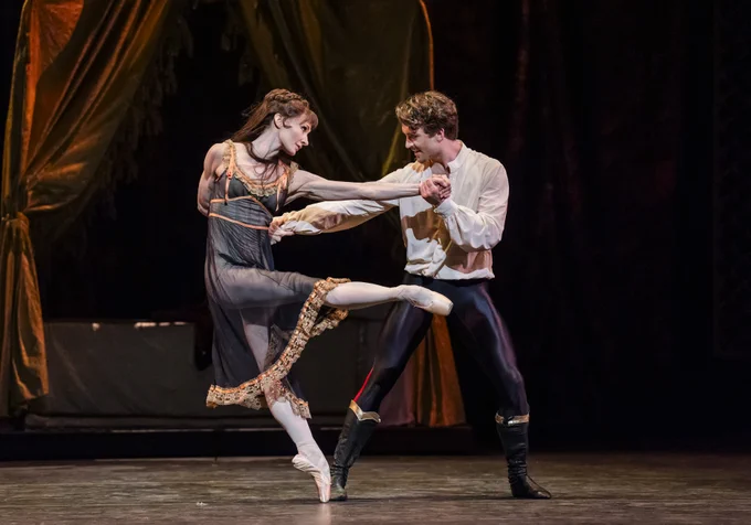 Two ballet dancers perform on stage. One is poised on pointe, while the other holds their hand. They wear period-style costumes. A draped backdrop and muted lighting create a dramatic setting.