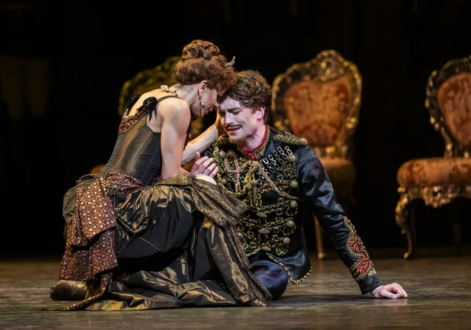 Two performers are on stage, one kneeling and the other leaning over, both in elaborate historical costumes. The backdrop includes ornate chairs, adding to the theatrical setting.