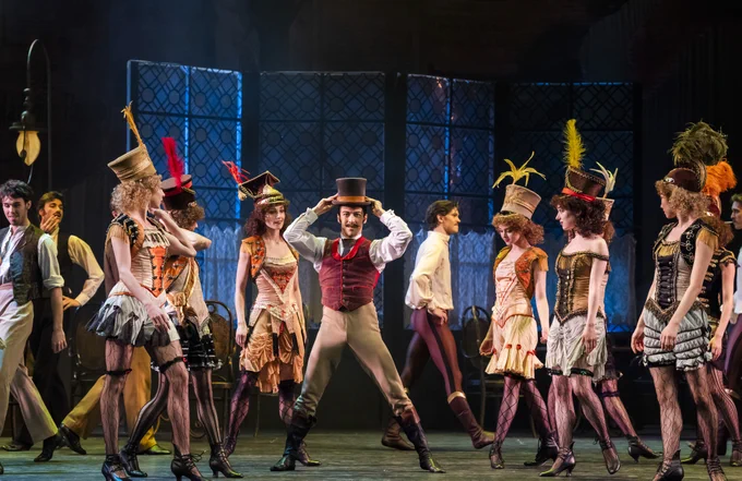 A theatrical performance featuring a group of performers in elaborate costumes with top hats and feathers, set on a stage with a vintage backdrop.