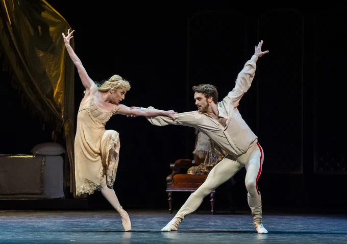 Ballet dancers perform an expressive, graceful duet on stage, likely depicting a scene from a classical ballet. One dancer extends an arm towards the other, creating a dynamic pose. The stage is minimally set, with a decorative backdrop and furniture pieces, enhancing the theatrical atmosphere.