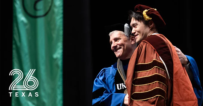 Two people in academic regalia are embracing on a stage, appearing to celebrate. One is wearing a blue gown with the letters "UK" visible, and the other is in a maroon gown with a cap. A green banner and the logo "26 TEXAS" are visible on the left.