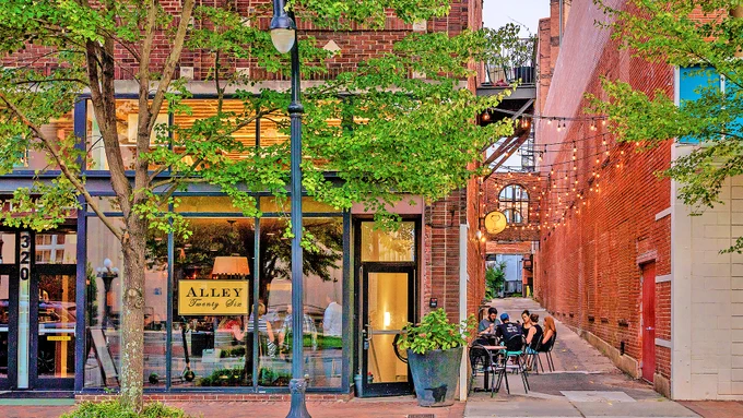 A cozy brick alleyway with string lights, outdoor seating, and people dining beside a storefront labeled "Alley Twenty Six" on a tree-lined city street.