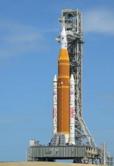 NASA\u2019s Artemis II Space Launch System (SLS) rocket and Orion spacecraft are seen atop a mobile launcher at Launch Complex 39B, Monday, March 30, 2026, at NASA\u2019s Kennedy Space Center in Florida. The orange core stage of the rocket stands between two white solid rocket boosters and is topped with the white Orion spacecraft. The rocket stands in front of the trussed structure of the mobile launcher. The sky is blue and cloudless in the background. Credit: NASA/Bill Ingalls