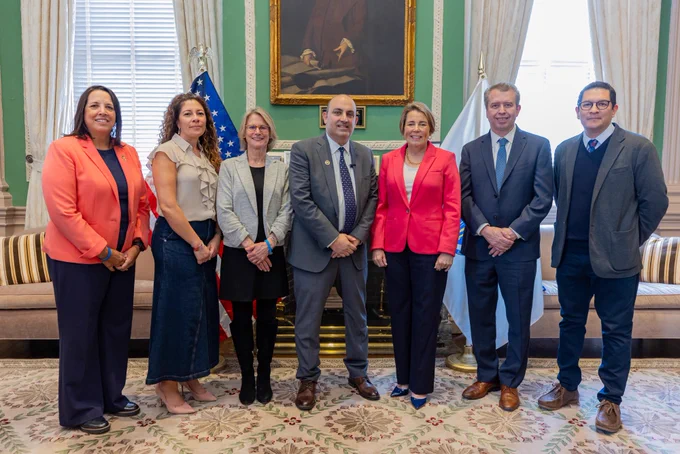 Education Secretary Steve Zrike in a gray suit stands with Governor Healey wearing a pink blazer in the State House’s Ceremonial Room. The two are flanked by Lieutenant Governor Kim Driscoll and state education officials posing for a group photo. The U.S. and Massachusetts flags and a framed portrait hang in the background.