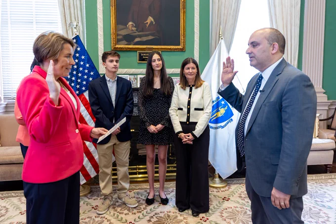 A formal swearing-in ceremony inside the Ceremonial Room inside the Massachusetts State House. Governor Healey in a bright pink blazer administers the oath to Education Secretary Steve Zrike in a gray suit, both raising their right hands. Zrike’s wife and two children stand behind them watching. U.S. and Massachusetts flags and a framed portrait hang in the background.