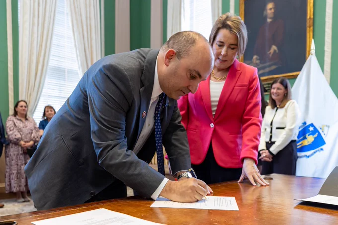 Education Secretary Steve Zrike in a gray suit signs official documents on a wooden desk after being sworn in, while Governor Healey in a pink blazer stands beside him observing. Several people watch from the background, with a Massachusetts state flag and framed artwork visible in the room.