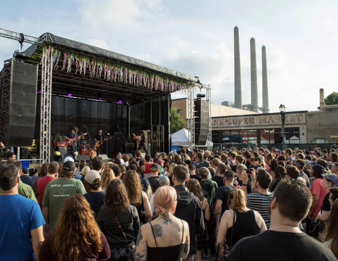 A band plays on an outdoor stage for a large crowd. The stage has floral decorations, and \"text@1\" SUPER MARKET\" is visible on a nearby building. Three tall smokestacks rise in the background.