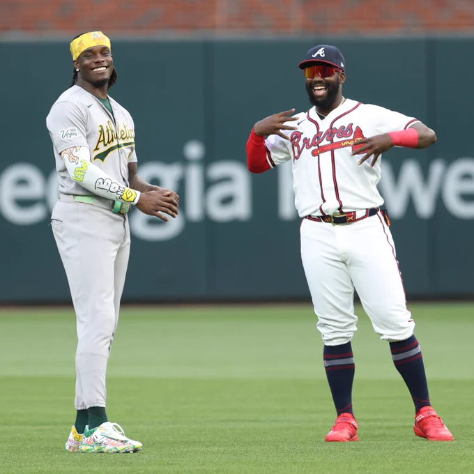Lawrence Butler and Michael Harris II smile in the outfield before Monday night's game at Truist Park. Butler wears a gray Athletics uniform with green lettering and yellow trim. Harris II wears a white Braves uniform with red lettering and navy trim.
(Photo: Cole Carter, Major League Baseball)
