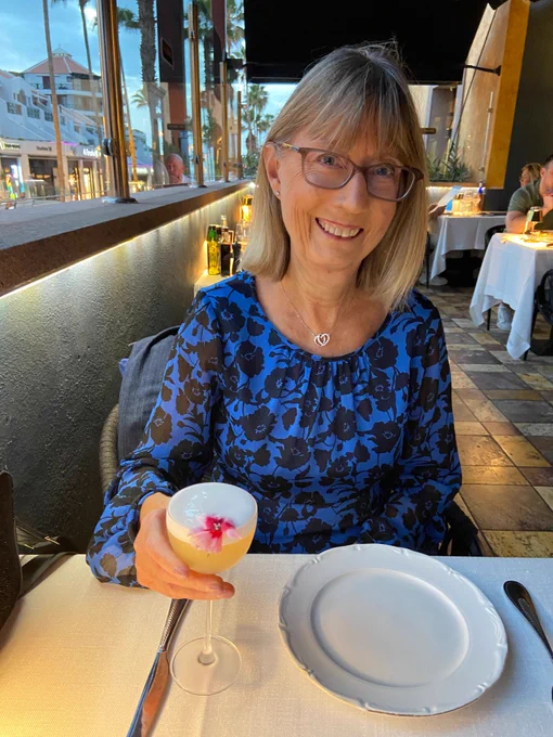 Janelle is a white woman in her late fifties with short, light hair and a fringe. She wears glasses and a long sleeved, dark blue top with black flowers on it. She's at a restaurant with a fruity looking cocktail. 