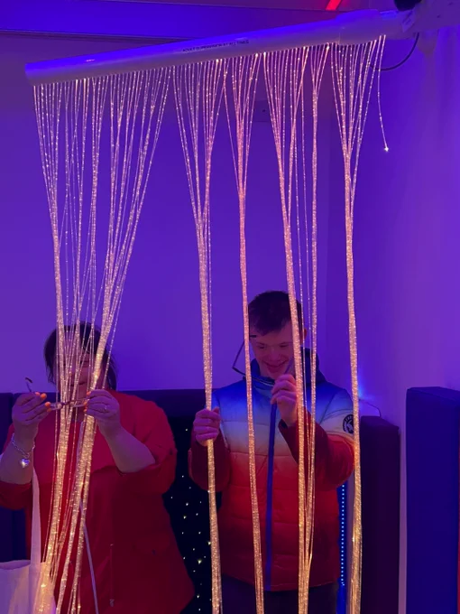 Two people are interacting with illuminated fibre optic strands hanging from above in a sensory room with soft purple lighting.