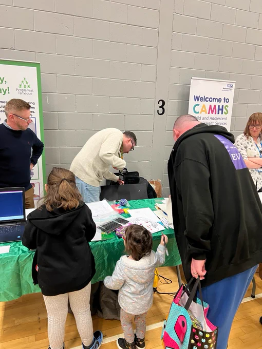 A group of people are gathered around a table at an indoor event. There are informational banners from "CAMHS" and "Together for Children" visible in the background. 