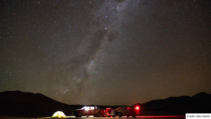 Brightly lit tents and vehicles under a clear night sky filled with stars above mountainous terrain.