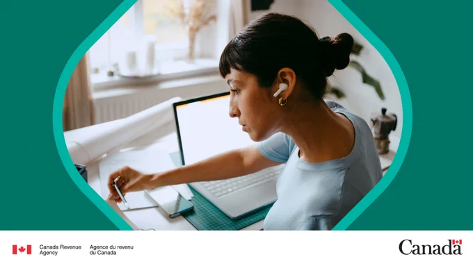 A person sits at a desk in front of an open laptop. They’re wearing earbuds and holding a pencil while looking at some papers.