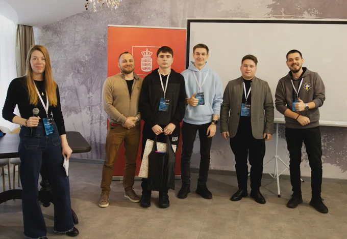 Six participants stand on stage during the award ceremony for the winners of the Cyber Mykolaivshchyna youth quest, holding gift bags and wearing event badges; a Government of Denmark banner is visible in the background.