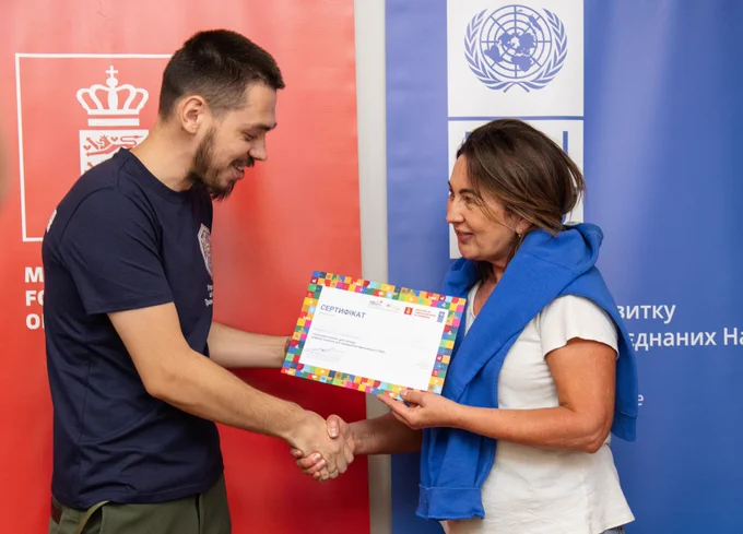 A male participant of a comprehensive training of trainers' programme on artificial intelligence (AI) smiles as he receives a certificate of successful training completion from a UNDP staff member. The two shake hands against the backdrop of UNDP and Government of Denmark banners.