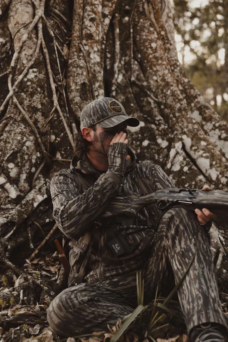 A person in camouflage attire, including a cap and full-face covering, is seated against a large tree with intricate roots. They are holding a hunting shotgun and appear to be in a forest setting, surrounded by fallen leaves and grass.
