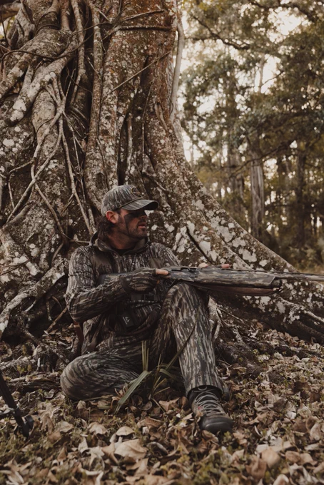 A person in camouflage clothing sits on the ground in a forest, leaning against a large tree with extensive roots. They are holding a shotgun and appear to be attentively observing their surroundings. The ground is covered in dry leaves, and tall trees are visible in the background.