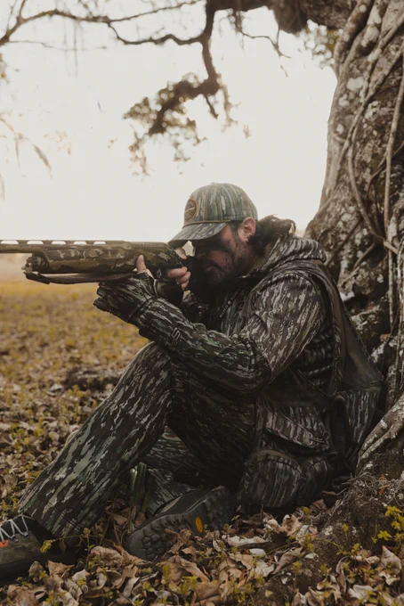 An individual in camouflage clothing is sitting against a tree, aiming a camouflaged shotgun. The ground is covered with leaves, and there are tree branches overhead. The scene suggests a hunting setting.