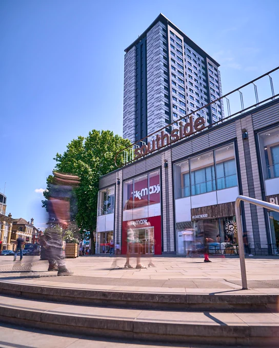 A tall building stands behind a shopping center called Southside, with a TK Maxx store visible. The scene includes blurred pedestrians and a bright, clear sky.