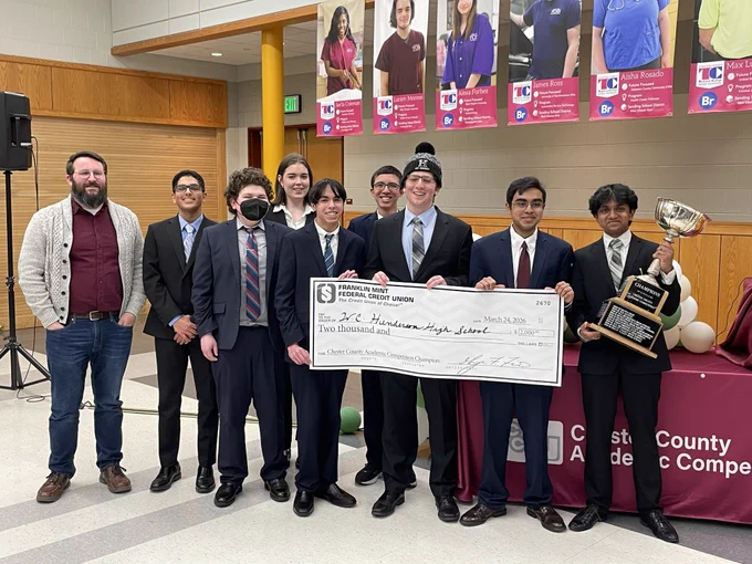 Group of young men in suits holding a large check and trophy at a career academy competition event indoors.