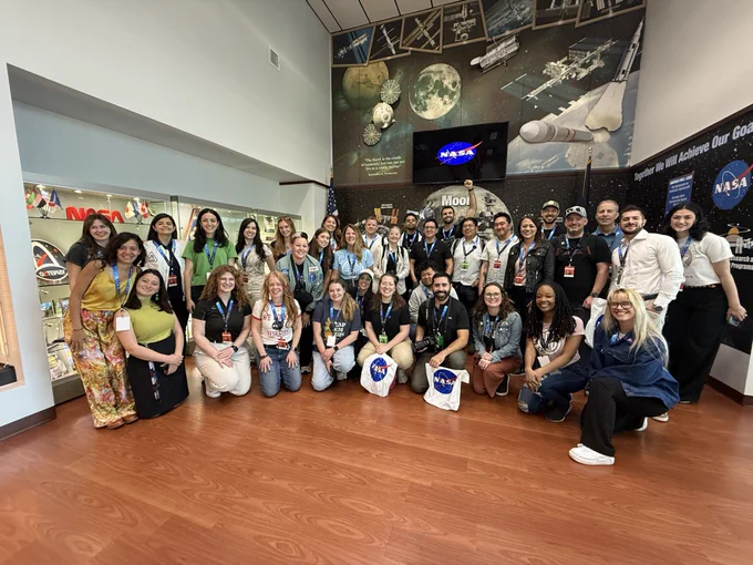 A group of approximately 30 people pose for a photo. Behind the group is a wall with a painted mural of the Moon and NASA logos.