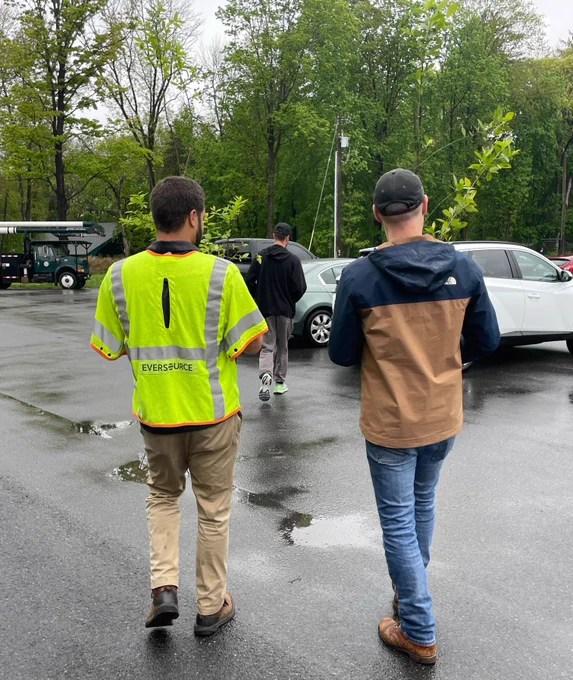 Eversource arborist wearing a bright yellow safety vest helps a customer carry trees through a parking lot.