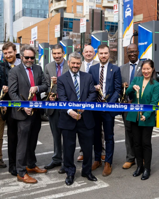 A group of city officials and partners stand outdoors holding ceremonial scissors as they cut a blue ribbon labeled "D O T Fast E V Charging in Flushing", marking the opening of a new electric vehicle fast charging station. 