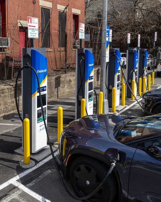 A row of blue and white fast charging stations lines a curbside lot, with a car plugged in and charging, protected by yellow bollards. 
