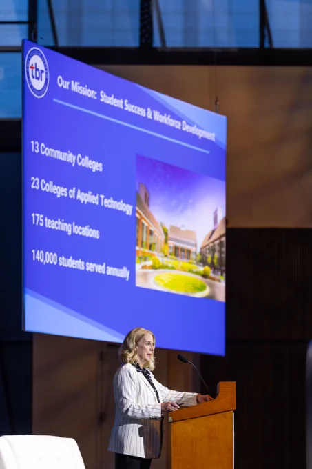 A person, Chancellor Flora Tydings, is speaking at a podium in front of a screen displaying a presentation, titled "Our Mission: Student Success & Workforce Development." It includes numbers of community colleges, Tennessee colleges of applied technology, teaching locations, and students. The logo "TBR" is visible on the screen.